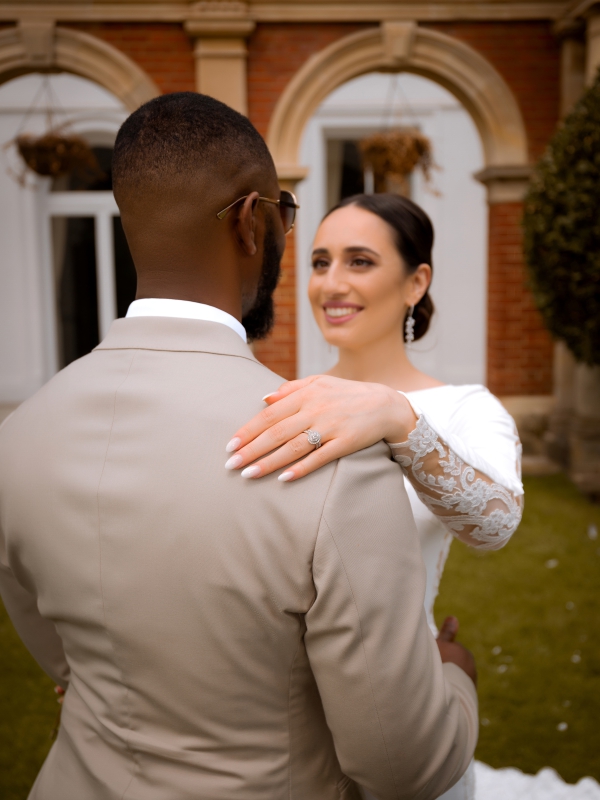 Close Up Of A Bride In A Long Sleeved White Gown Smiling At Her Groom, With Her Hand Resting On His Shoulder.